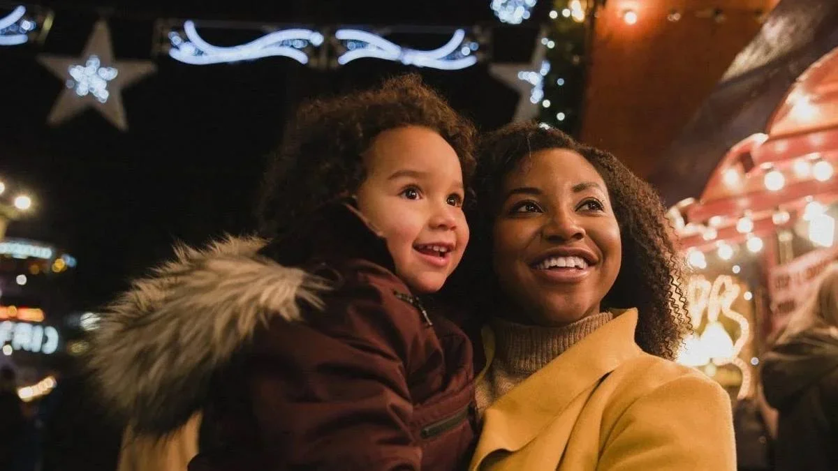 A close-up front-view shot of a woman holding her young daughter, they are both looking at bright Christmas lights at the Christmas markets on a cold night, the young girl is in awe.